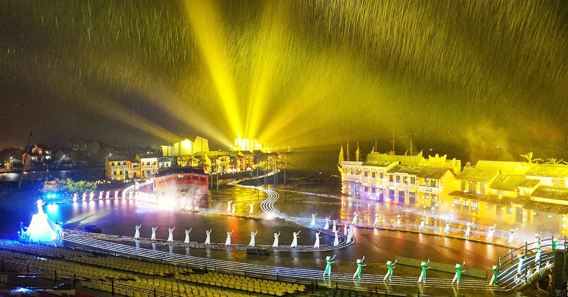 A night-time stage show with golden light beams over a lit riverfront set